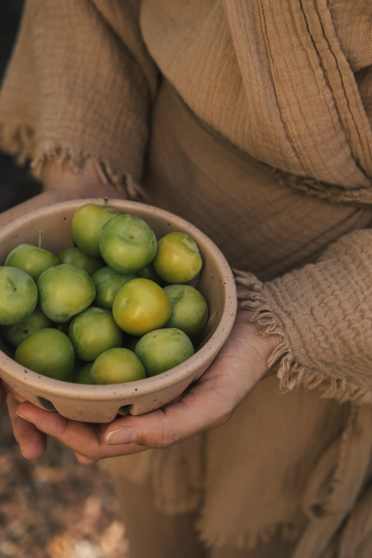 Hands in textured linen hold a speckled ceramic colander brimming with fresh limes.