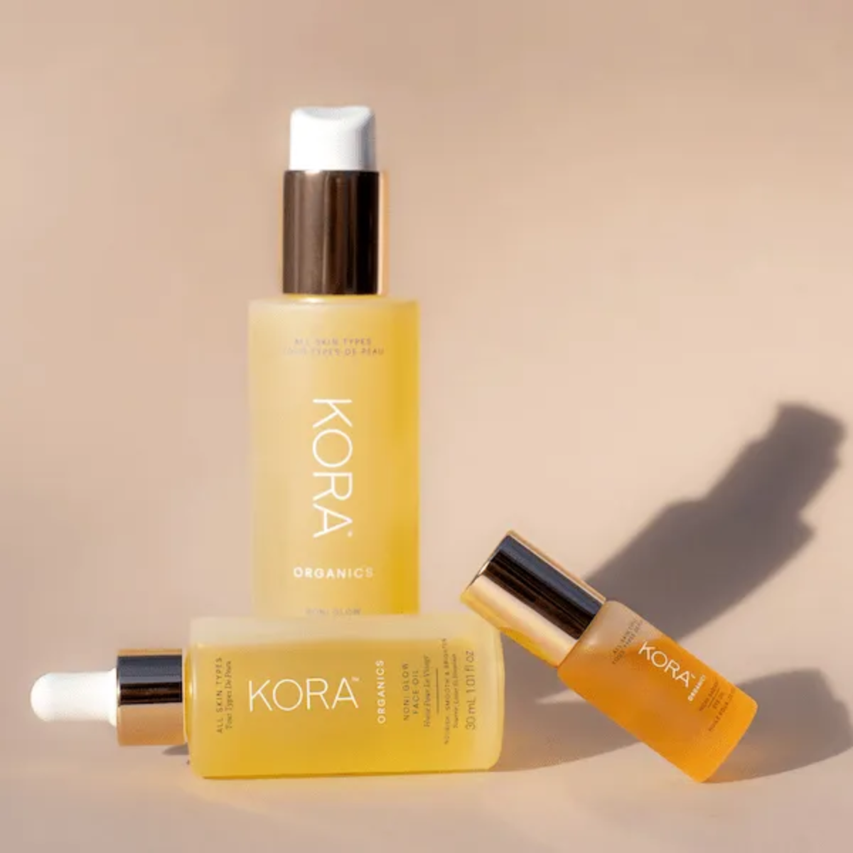 Three KORA Organics Noni Glow Face Oil bottles in various sizes arranged on neutral background.