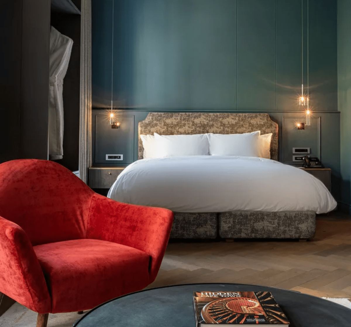 Modern boutique hotel room in Cape Town with a teal paneled wall, crisp white bed, red velvet armchair, and ambient pendant lighting.