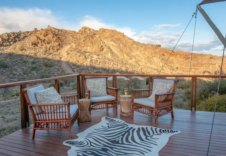Elevated wooden deck at Sanbona Wildlife Reserve with rattan armchairs, zebra-print rug and golden Karoo mountains glowing in late-afternoon sun.