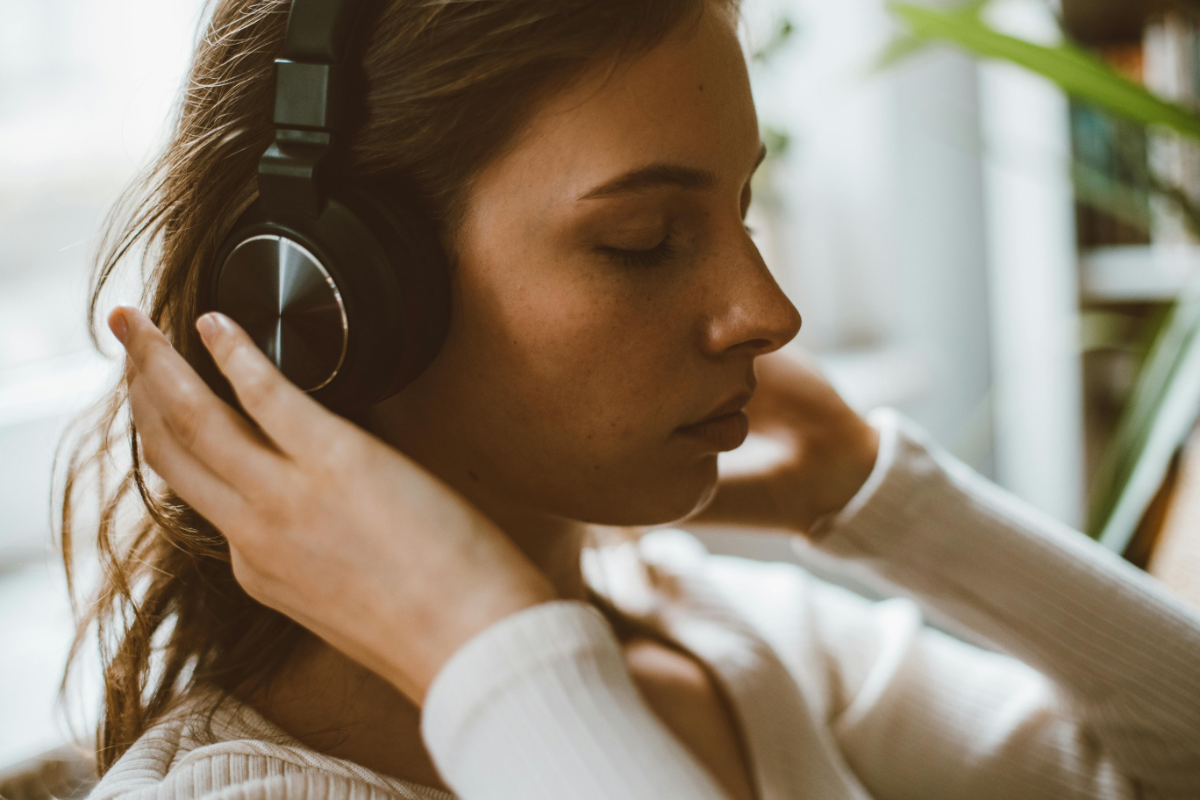 Close-up of a young woman indoors, eyes closed, gently adjusting black over-ear headphones while listening to music.