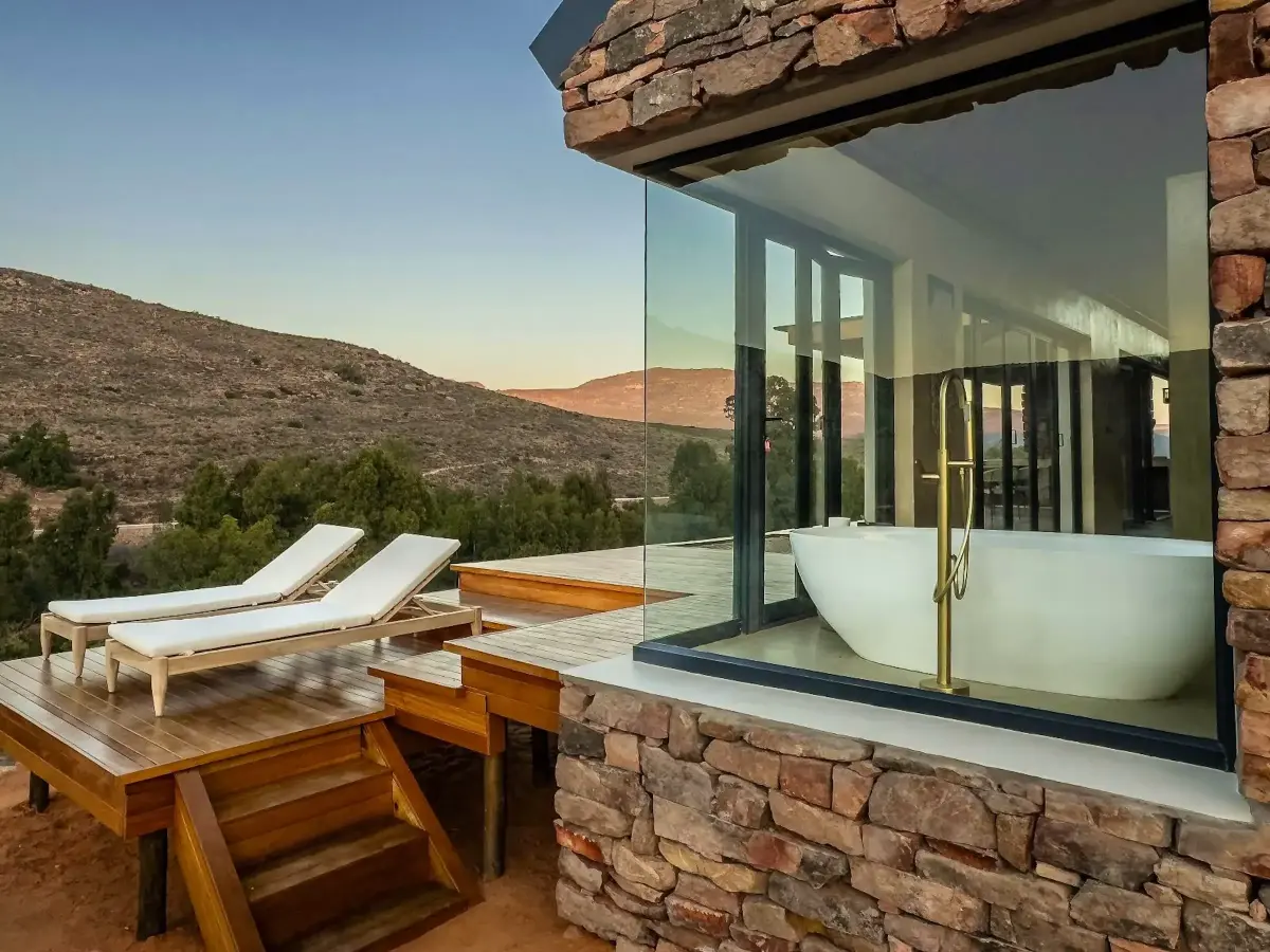 Stone-clad Ashante cabin with glass-walled soaking tub and timber sun-lounger deck overlooking the rugged Breede River mountain landscape.
