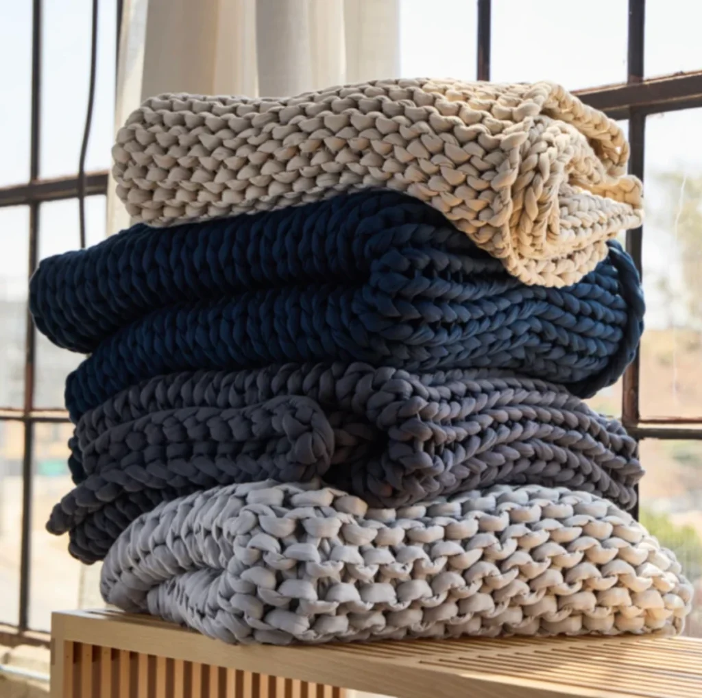 Avocado weighted blanket in chunky knit cotton stacked in neutral and deep tones by a window