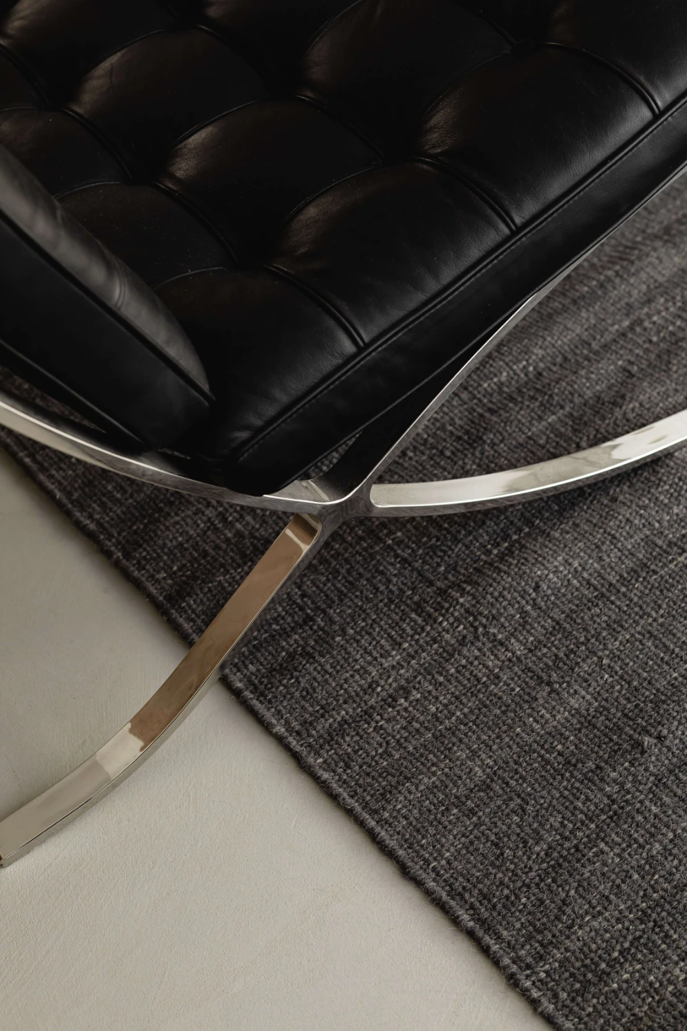 A tufted black leather Barcelona-style chair on a polished steel frame, resting on a charcoal wool rug.