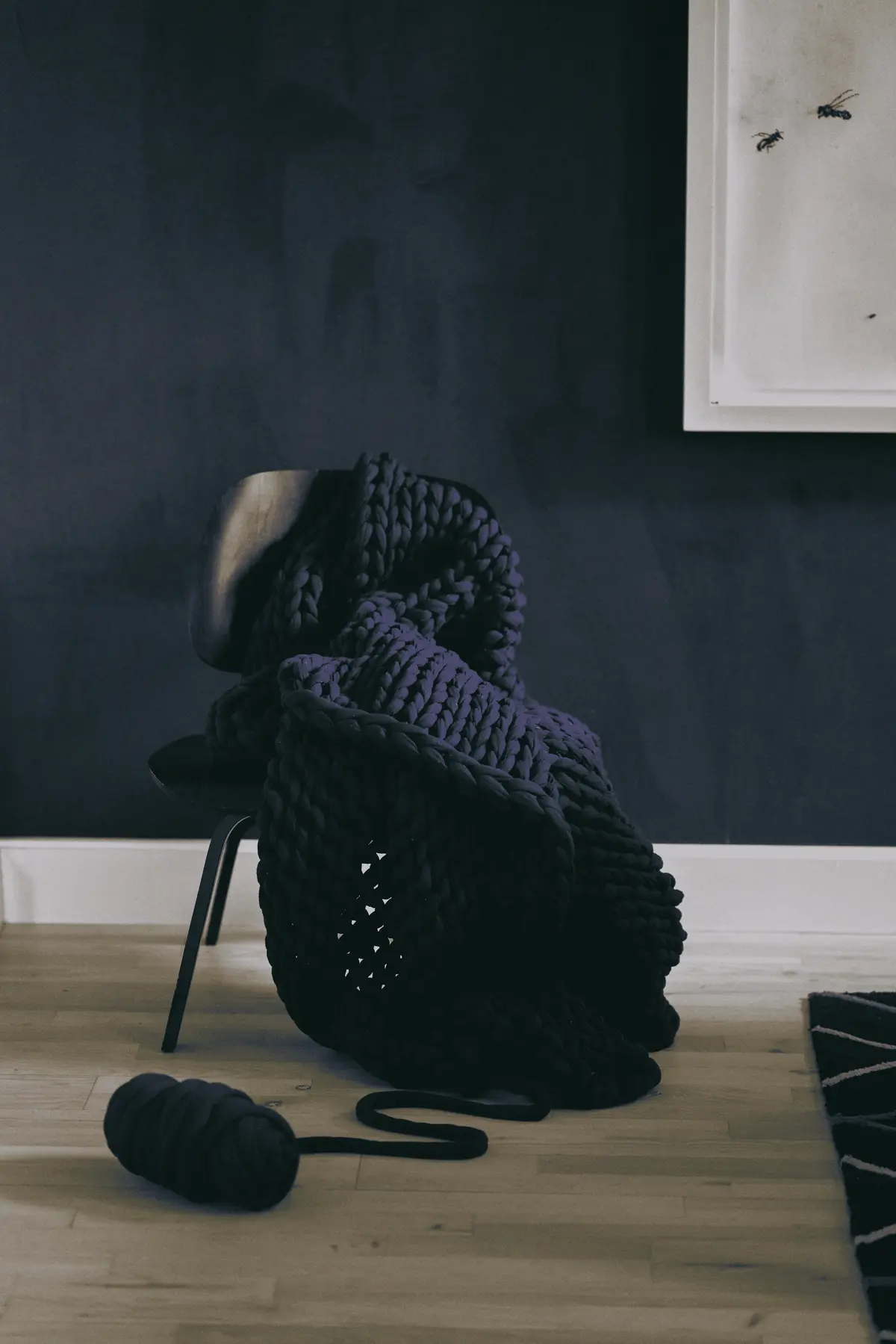 Navy blue chunky-knit weighted blanket draped over mid-century chair with matching yarn skein on hardwood floor.