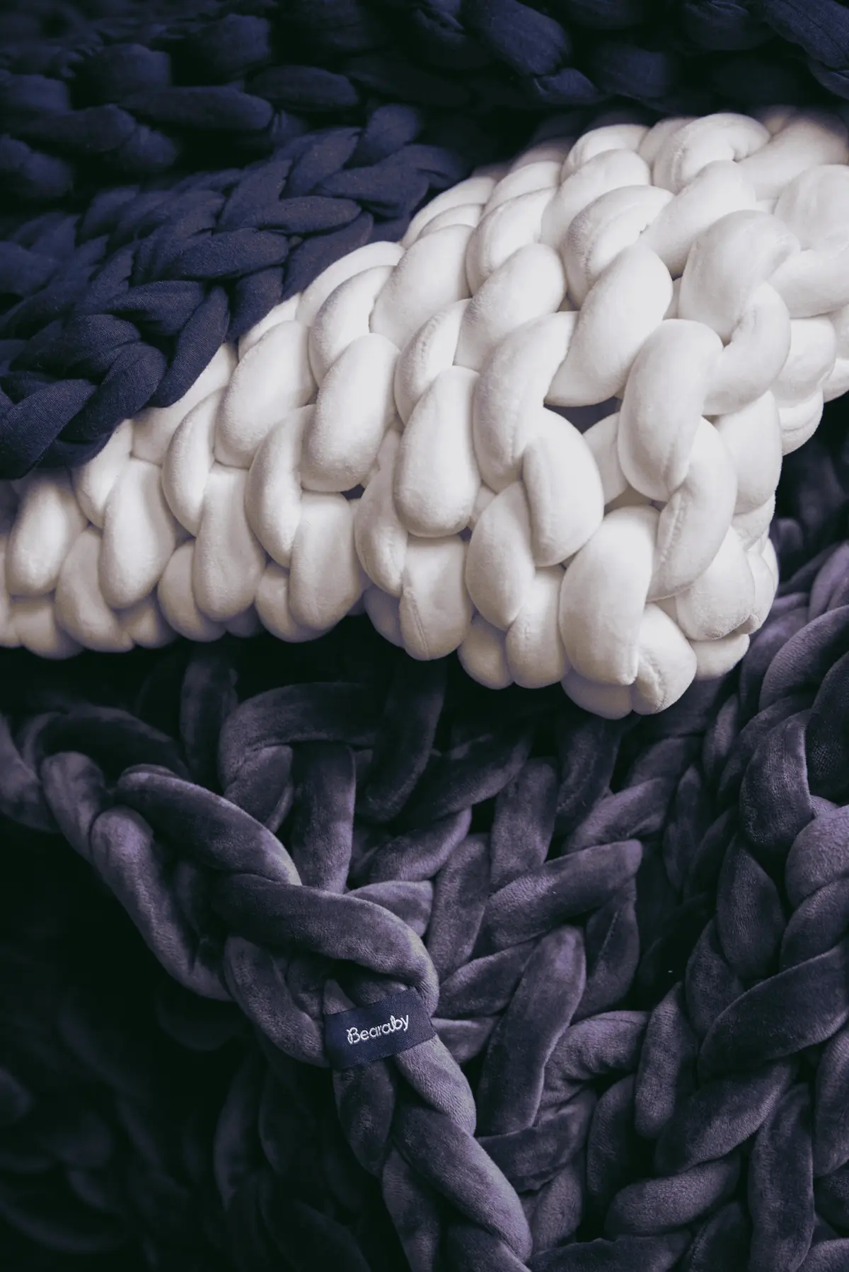 Stack of Bearaby chunky-knit weighted blankets in navy, ivory and plum displaying thick, plush loops.