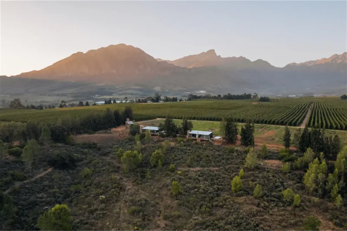 Sunset drone view of Bergsicht Outpost’s tin-roof cabins surrounded by orchards and the Witzenberg mountain range in South Africa’s Breede River Valley.