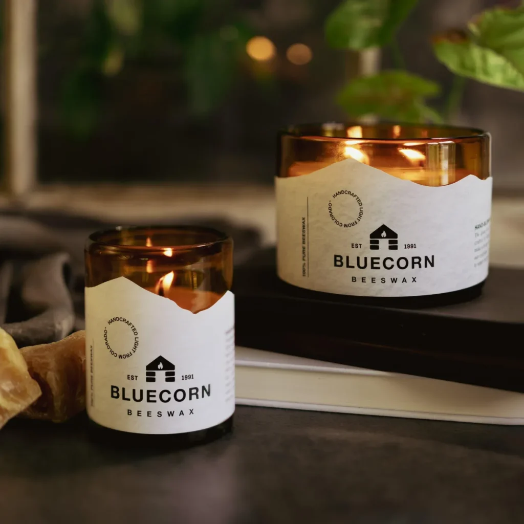 Two lit Bluecorn beeswax candles in amber glass jars with organic labels on stacked books