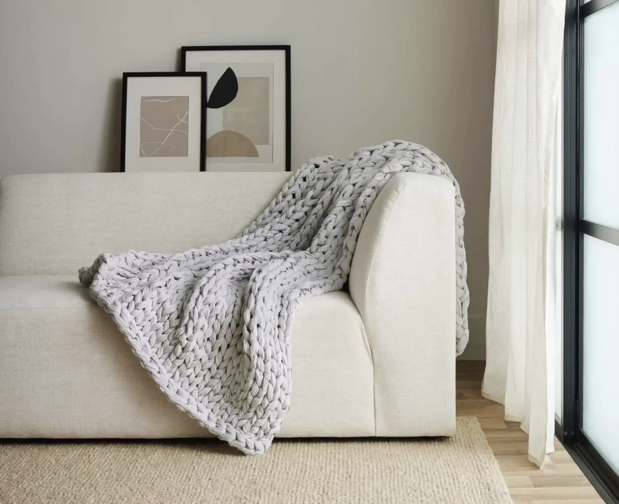 Light-grey chunky-knit weighted blanket casually draped over minimalist cream sofa with abstract art in background.