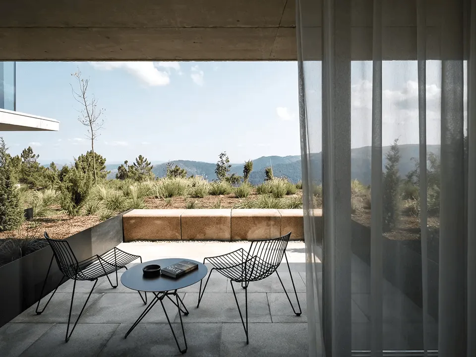 Minimalist balcony in northern Portugal, black wire chairs and coffee table facing rewilded rooftop garden and Douro valley hills.