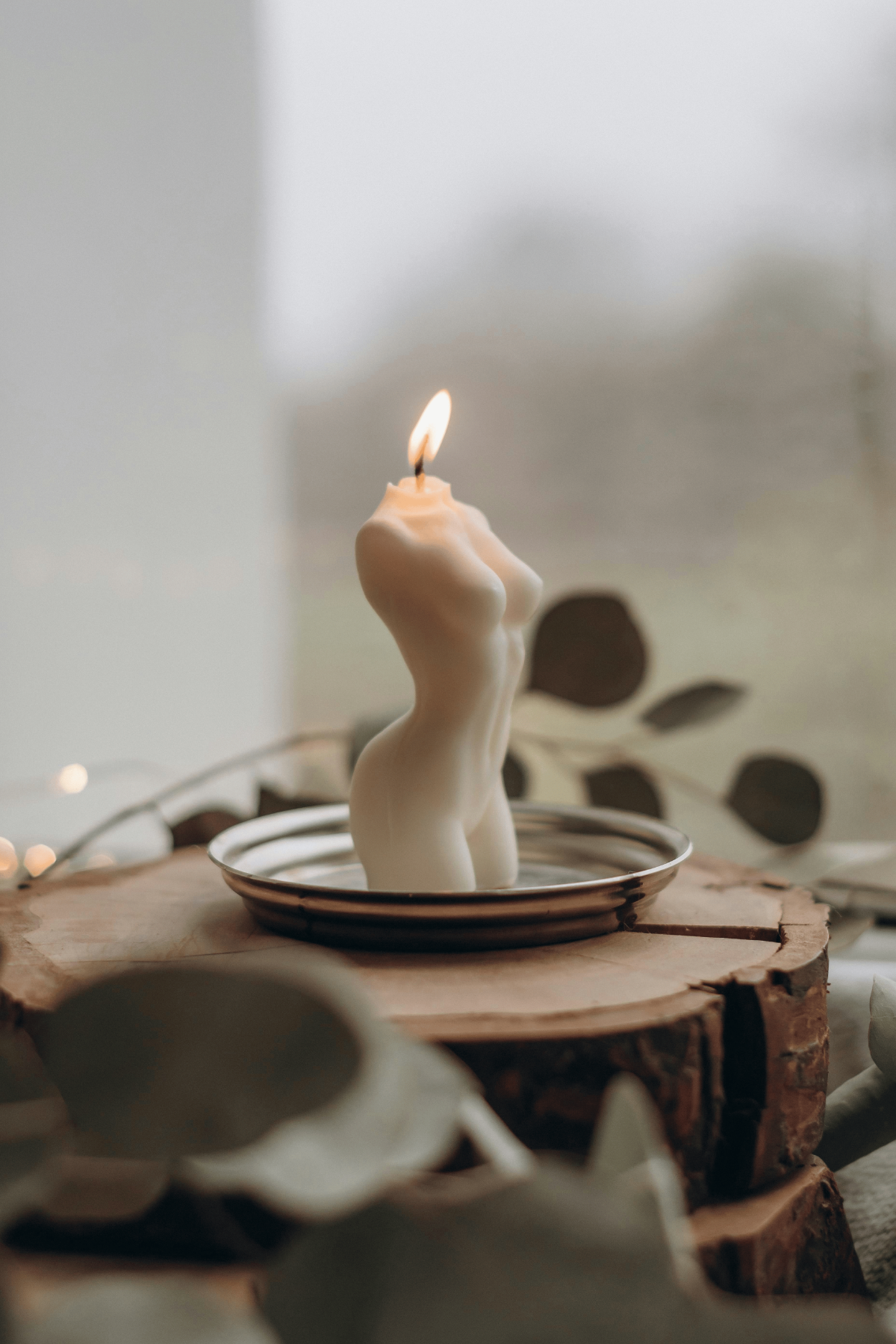 Lit female-torso soy candle on metal dish, styled with rustic wood and eucalyptus for elegant home-décor vignette.