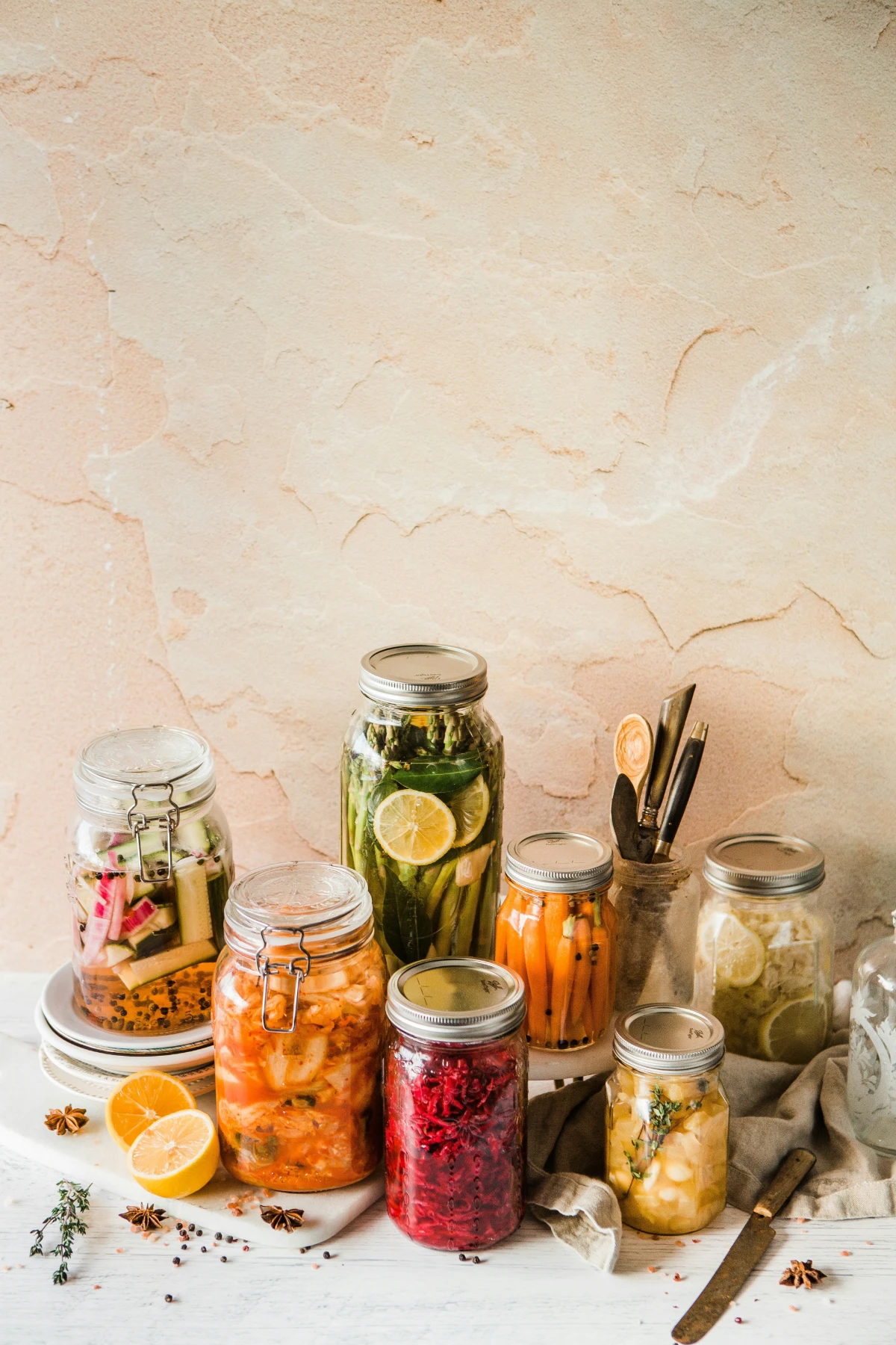 Assorted fermented vegetables in glass jars with lemon, spices, and herbs on a rustic kitchen surface.