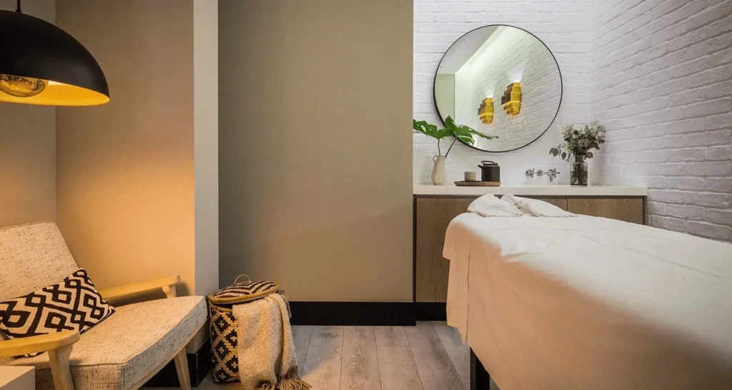 Cozy treatment room at The Lumiares: neutral tones, round mirror and single massage bed under soft amber pendant light