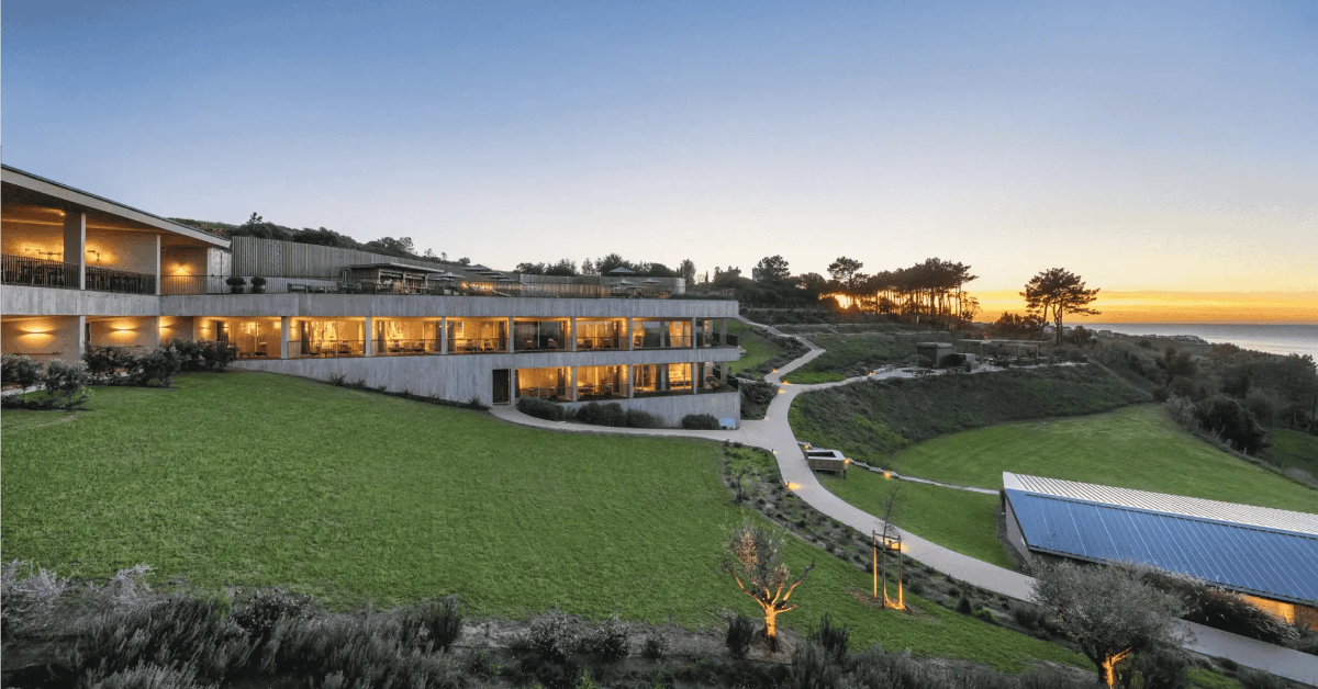 Tiered concrete-and-timber Immerso Hotel glowing at sunset on grassy cliff above Portugal’s Atlantic coast.