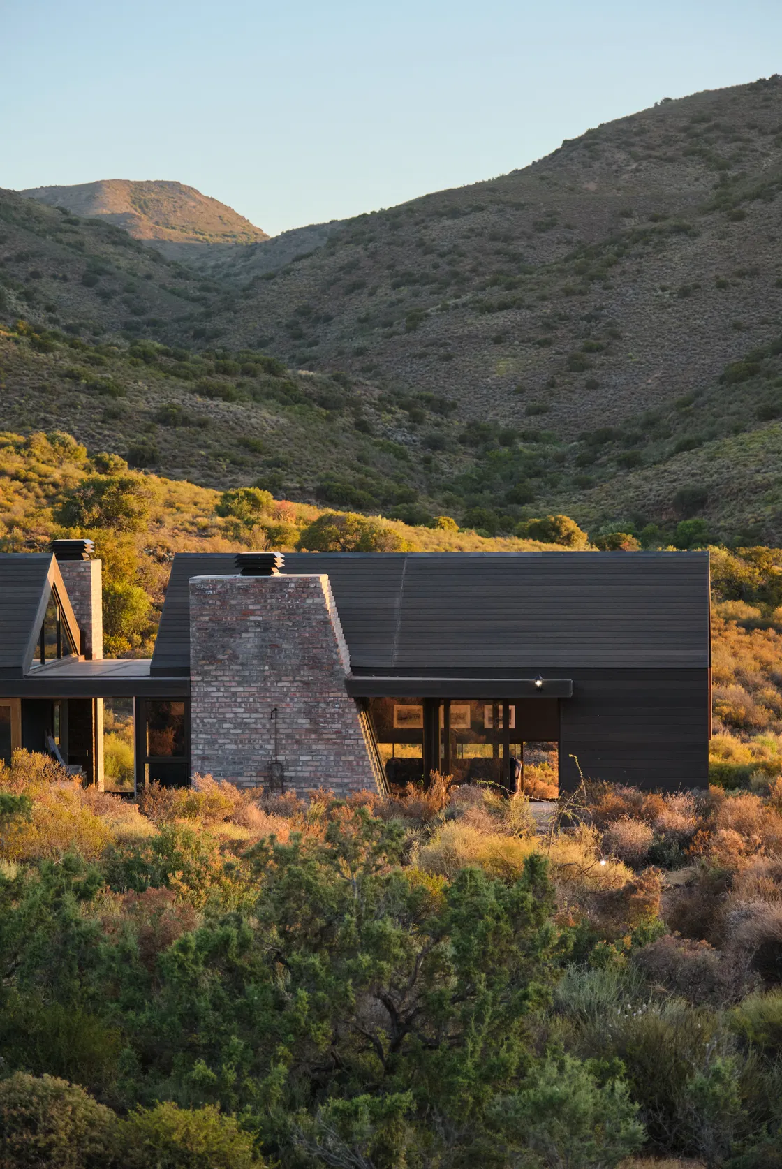 Dark-clad Barn House retreat with brick chimney, tucked into sunlit Karoo mountains and indigenous shrubland.