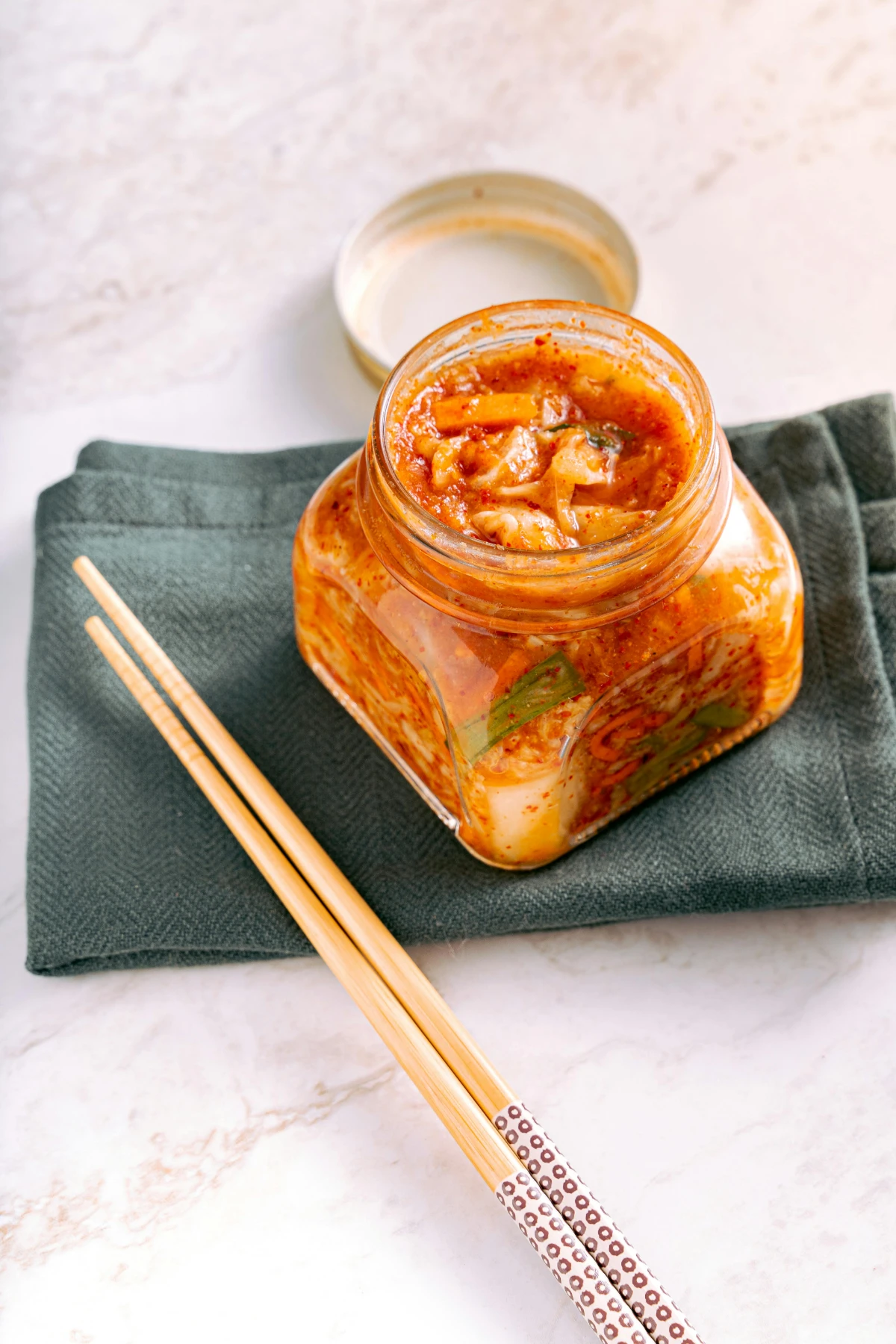 Open jar of kimchi with chopsticks on cloth, showcasing traditional fermented vegetables with vibrant texture.