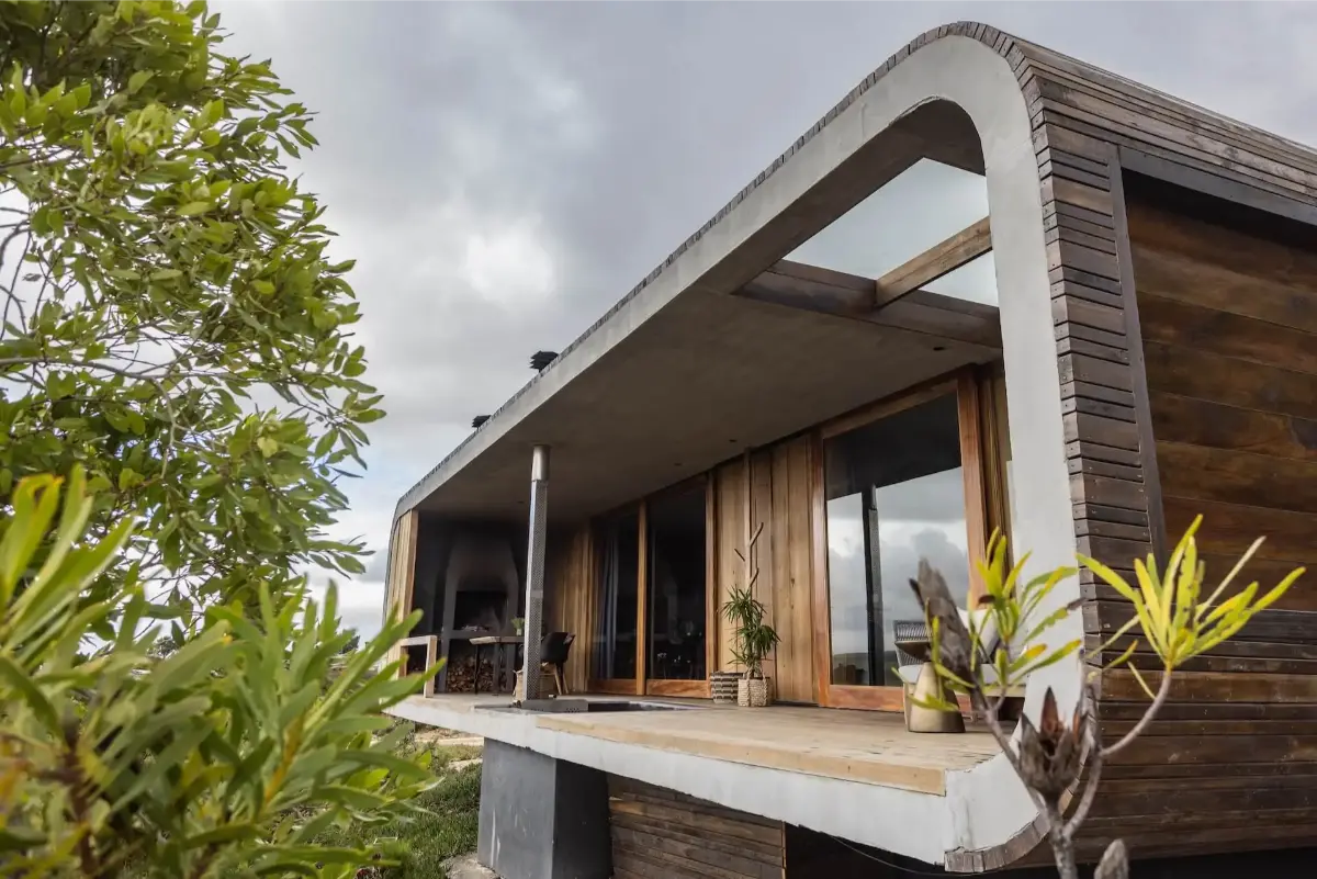 KolKol Pods’ Sunbird unit in Elgin: sculptural curved-concrete eco-pod with timber cladding, sheltered braai patio and fynbos foreground under moody skies.