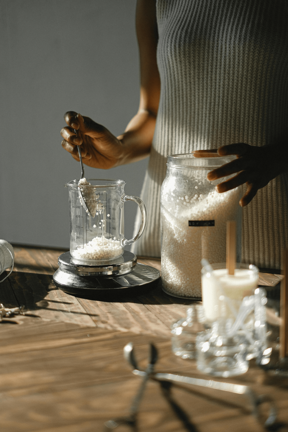 Home studio soy candle making: scooping wax pellets into beaker on digital scale, tools and finished candles in morning light.