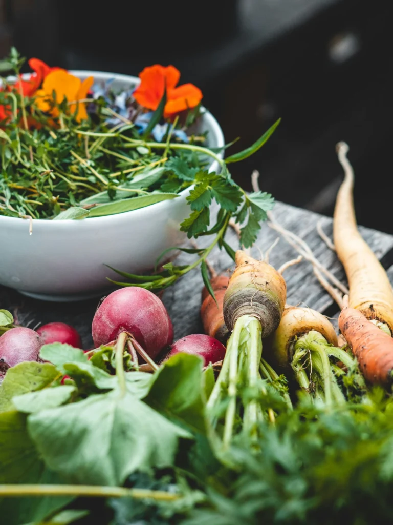 Seasonal root vegetables and fresh herbs beside a bowl of greens in a natural kitchen setting