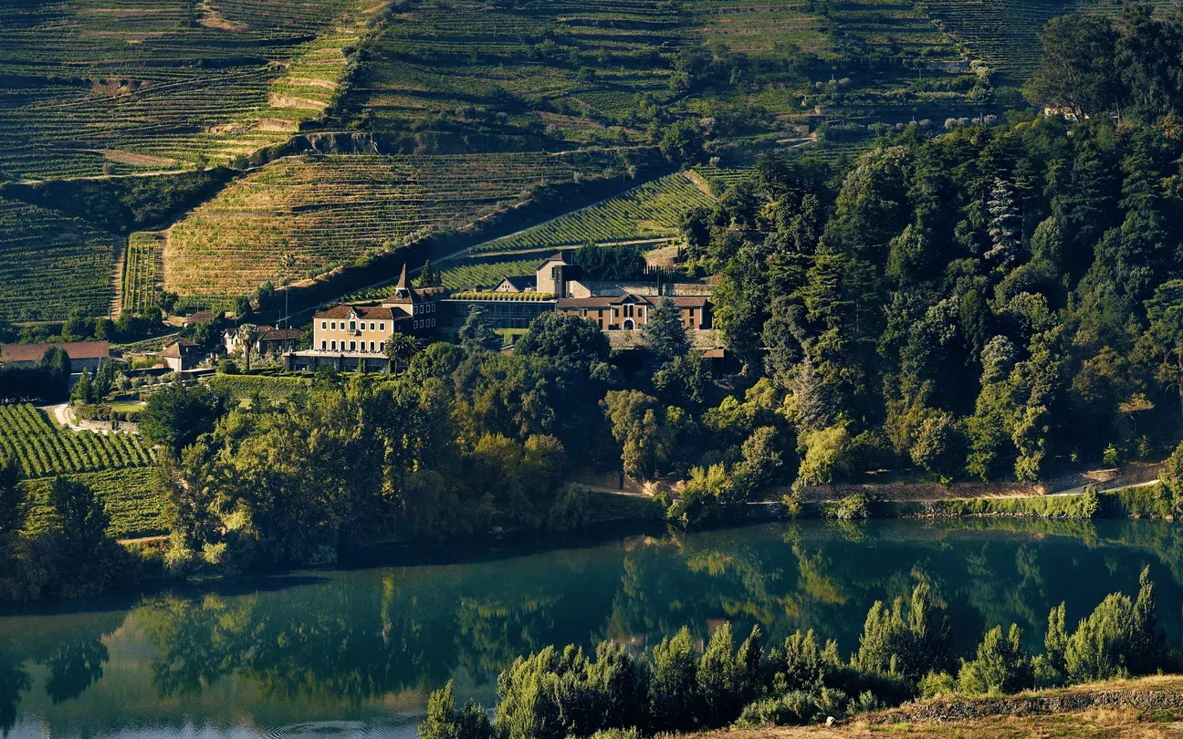 Six Senses Douro Valley estate nestled amid stepped vineyards and forest, reflected in the still river below.