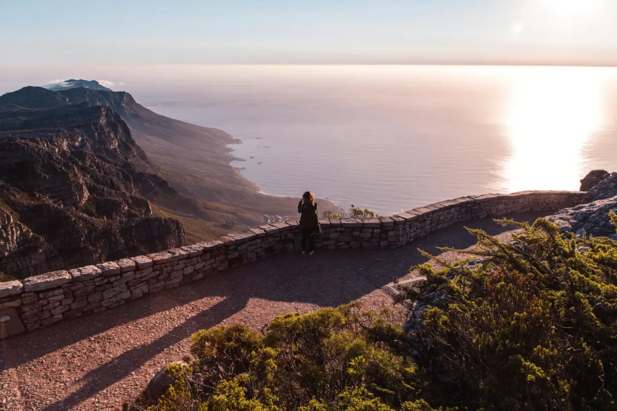 Visitor leans on summit lookout wall of Table Mountain, watching sun glitter across calm Atlantic horizon.