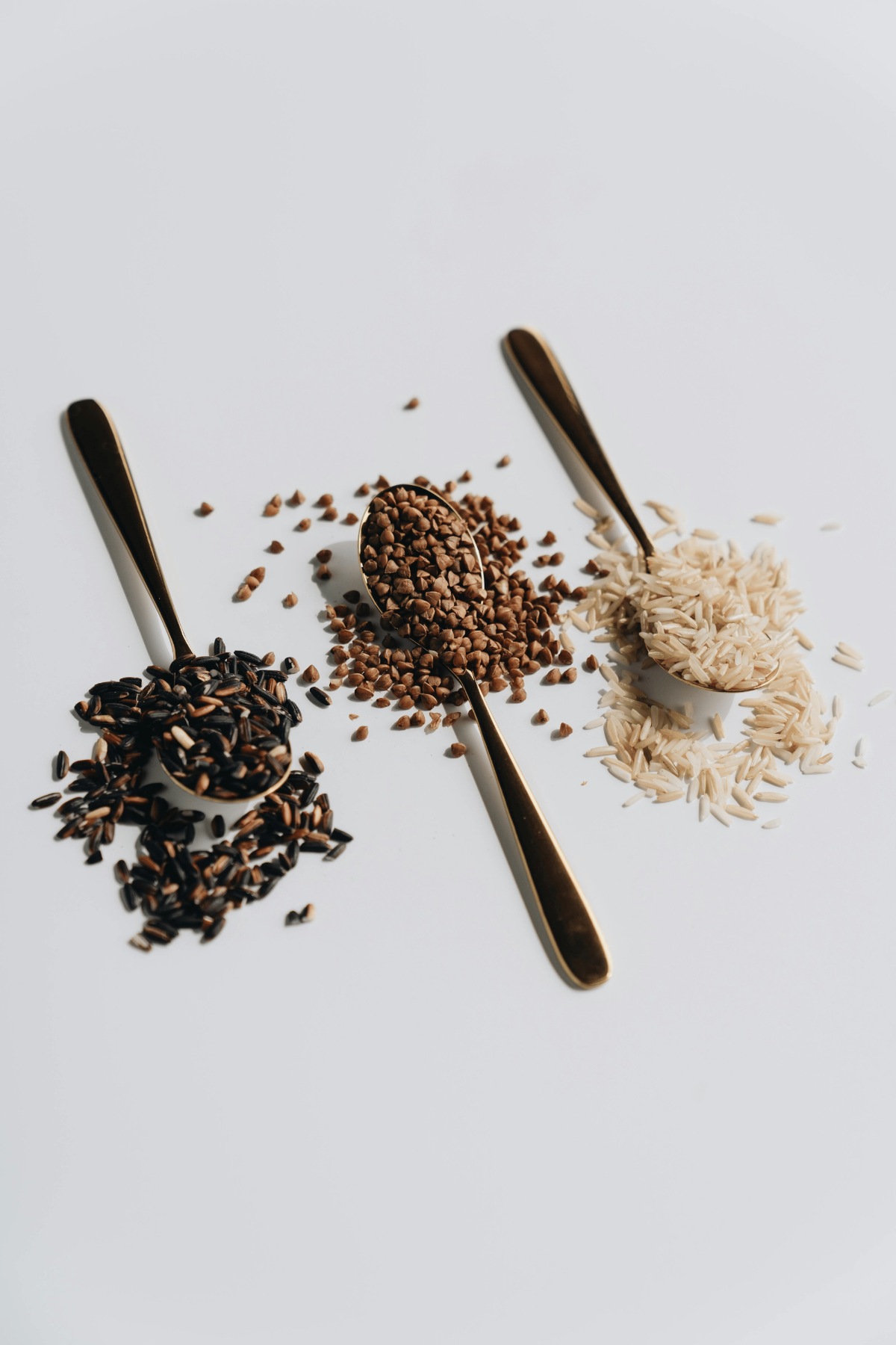Gold spoons displaying wild rice, buckwheat groats and long-grain white rice on minimal white surface to compare textures.