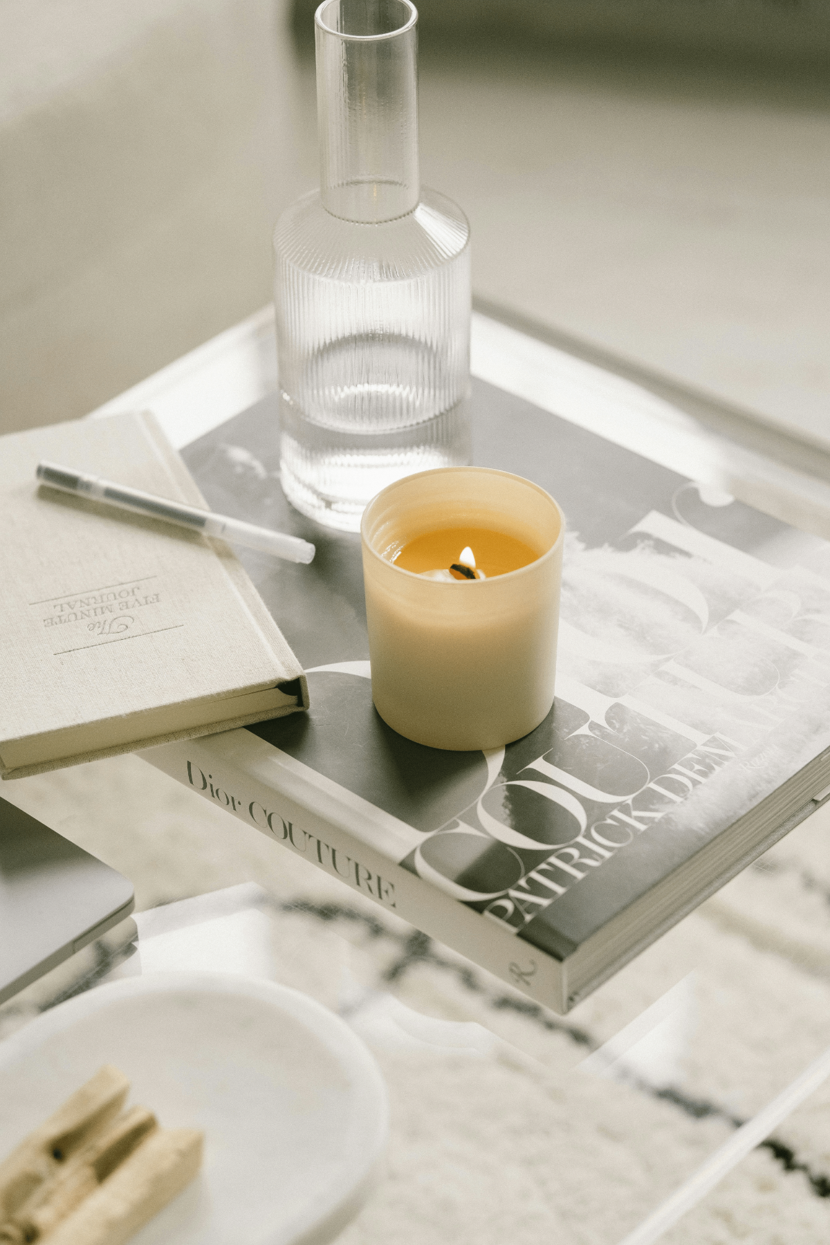 Neutral living-room flat lay with lit beige wood-wick candle on art books, glass carafe, and daily journal.