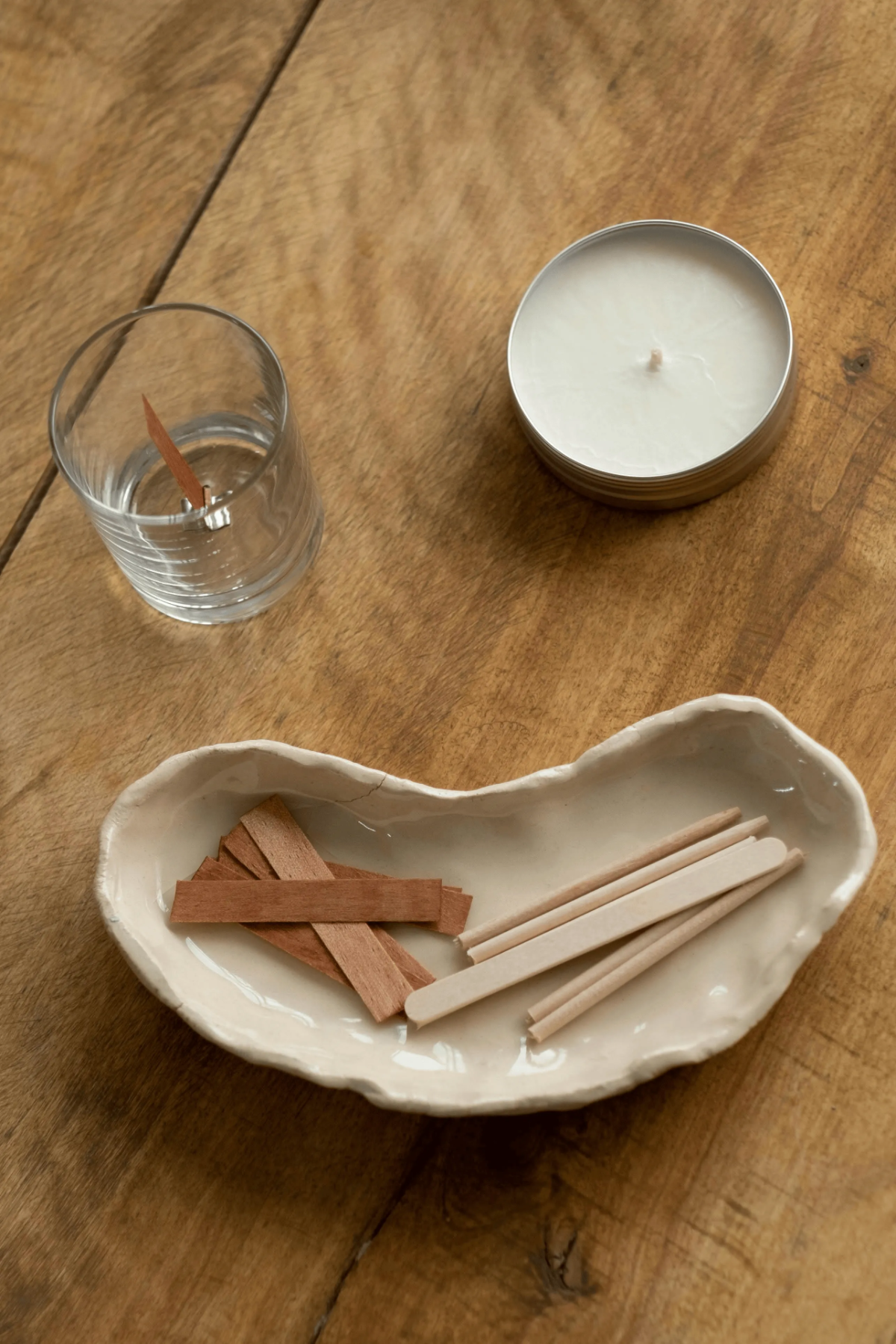 Flat lay DIY candle supplies: tin soy candle, glass tumbler with wood wick, fragrance strip, cedar sticks on rustic table.