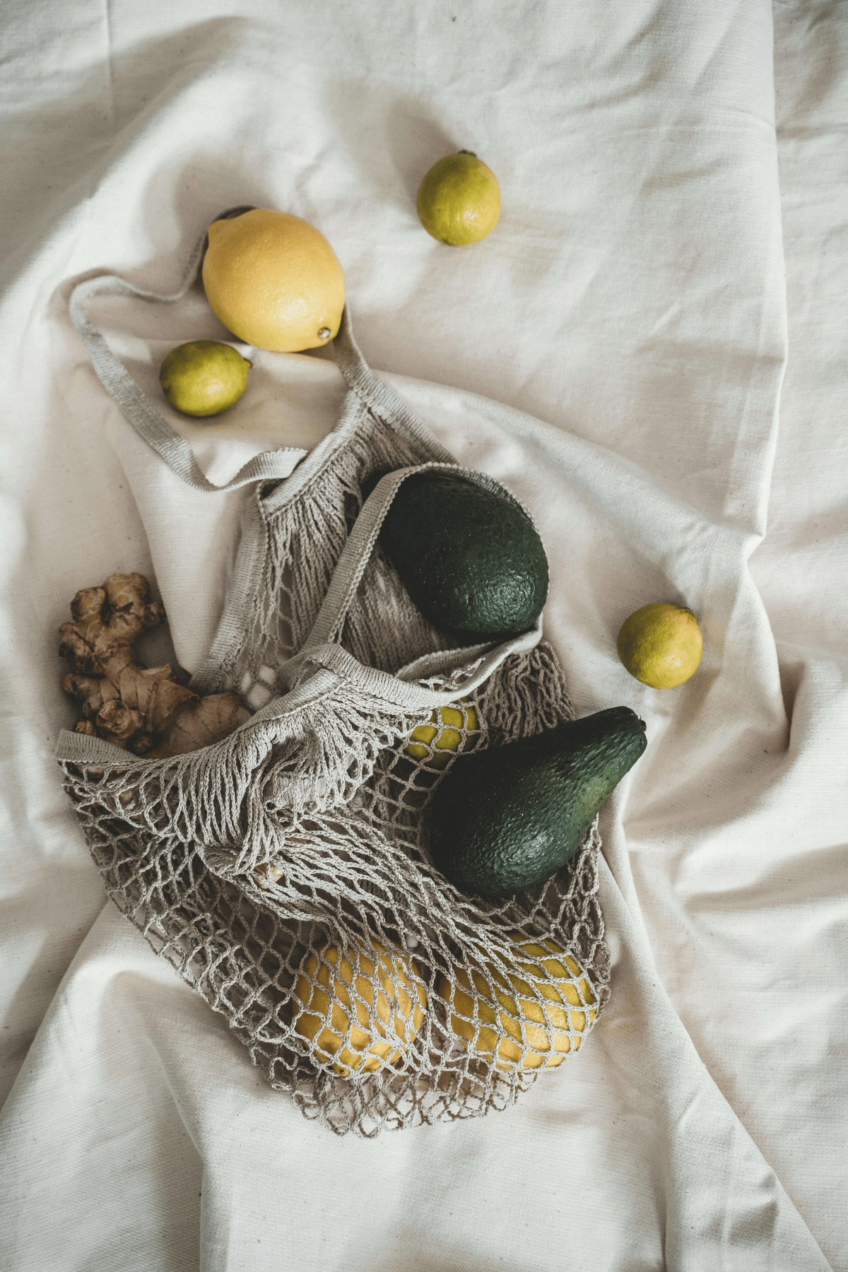 Reusable net bag with avocados, lemons, limes, and fresh ginger on cream linen.