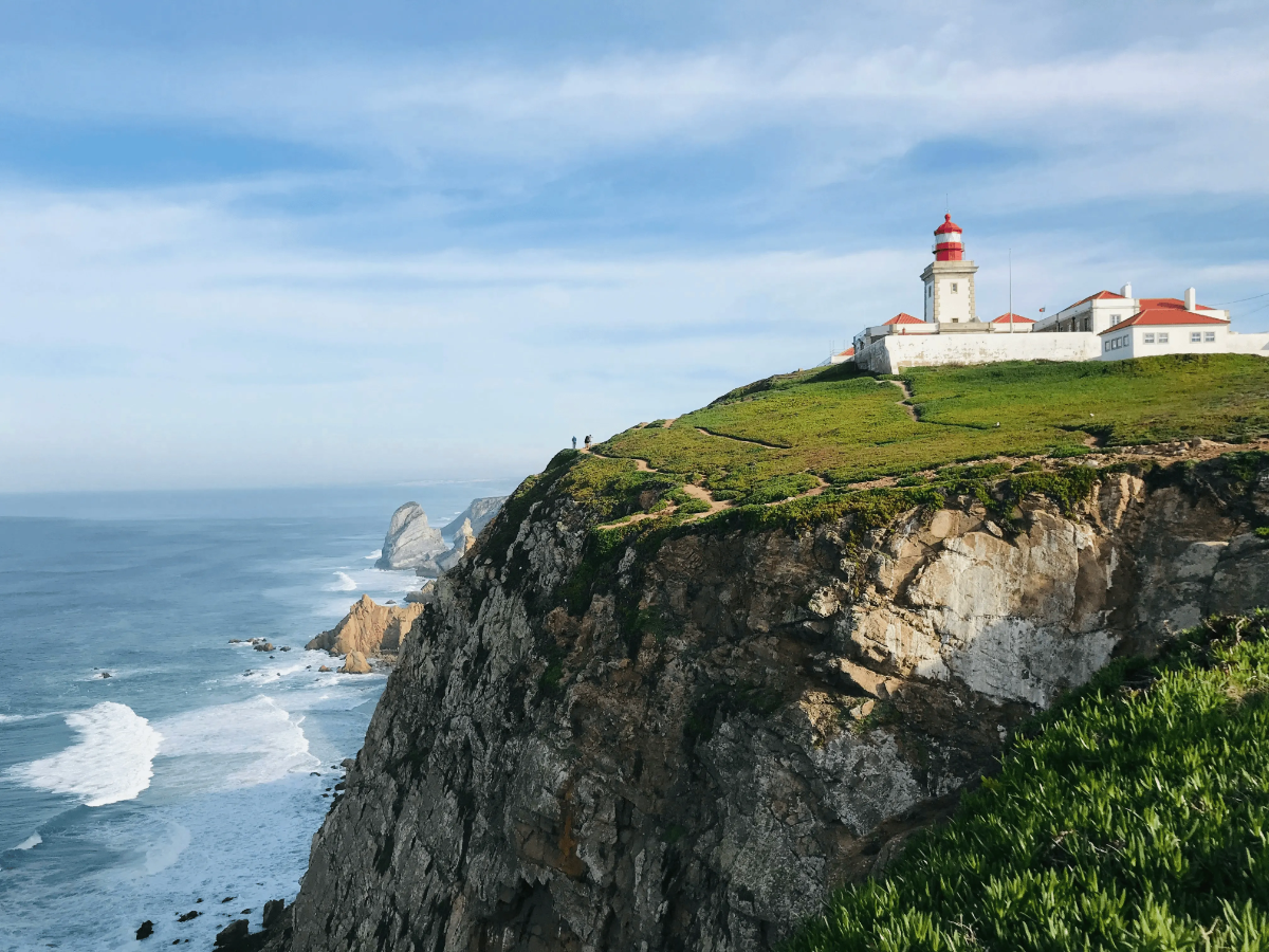 Red-topped Cabo da Roca lighthouse perched on a grassy Atlantic cliff high above crashing waves and rugged coastline.