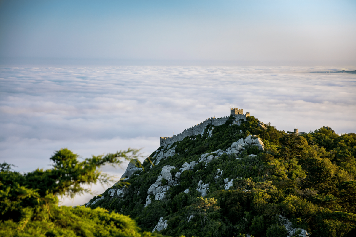 Castelo dos Mouros walls atop forested Sintra mountain, rising above a sea of clouds at sunrise.