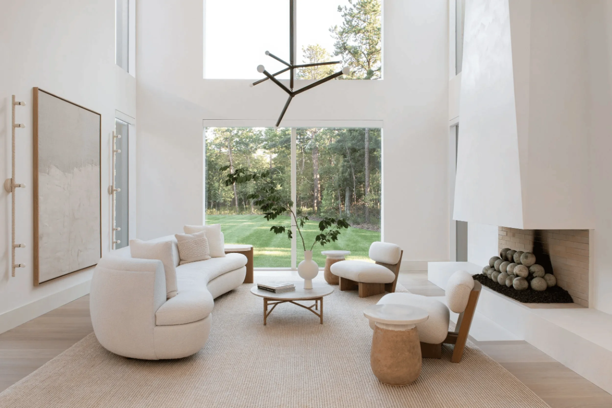 Chango & Co. modern Hamptons living room with white curved sofa, lounge chairs, sculptural fireplace, and large windows to a wooded lawn.