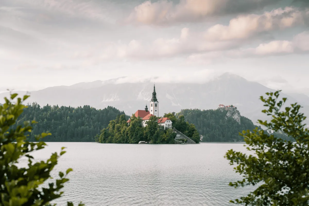 Misty morning view of Lake Bled’s island church framed by soft clouds, calm water and lush green shoreline.