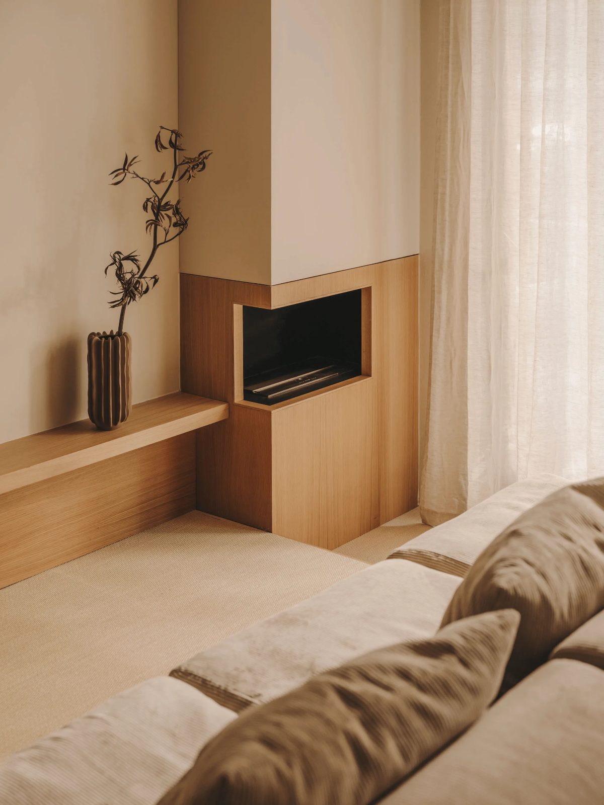 Warm minimal living room with wood-clad fireplace, linen curtains, ribbed vase and beige sofa by Miriam Barrio, inspired by Japanese kanso.