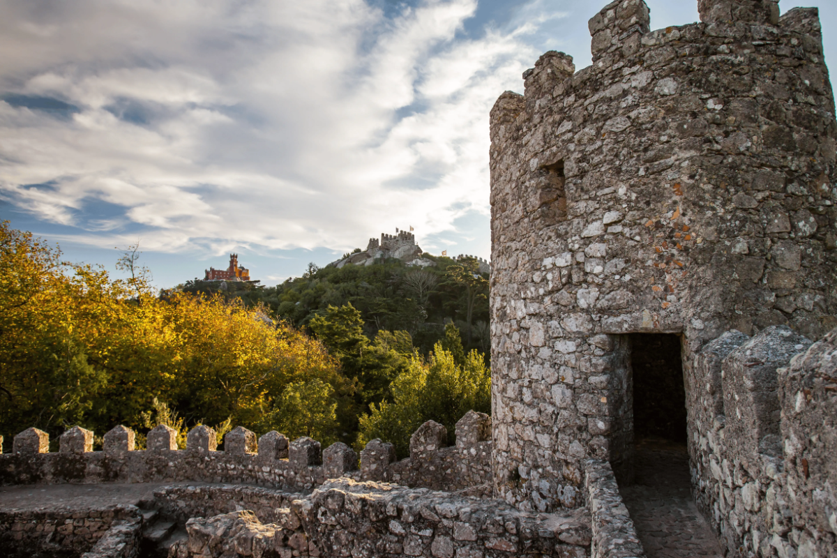 Stone battlement tower of Sintra’s Moorish Castle overlooking forested hills and distant Pena Palace under a dramatic sky.