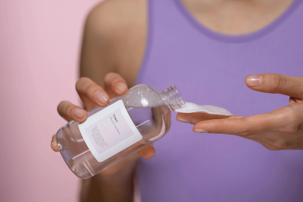 Woman in lavender top tips a clear bottle, saturating a cotton pad with cleansing liquid.