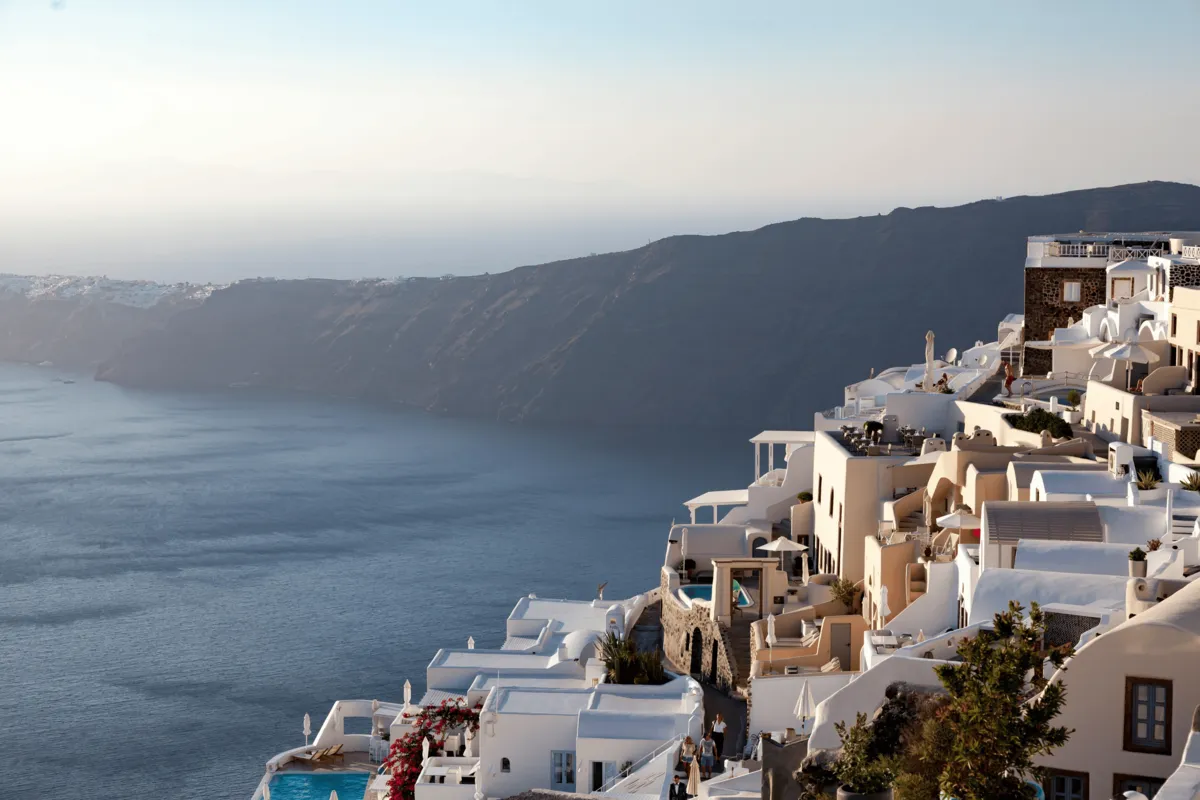 Whitewashed houses cascading down a sun-lit cliffside in Santorini, overlooking calm blue waters of the volcanic caldera at sunset.