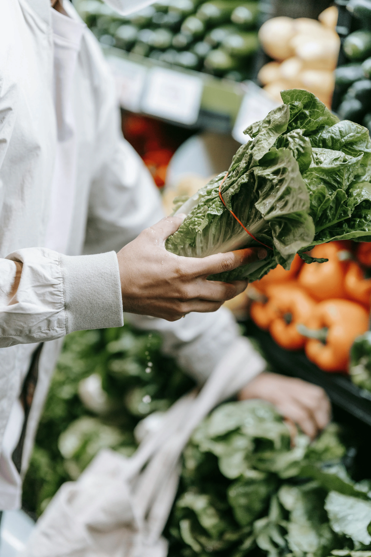 Shopper wearing mask selects bundle of crisp romaine lettuce in front of vibrant produce display at farmers’ market.