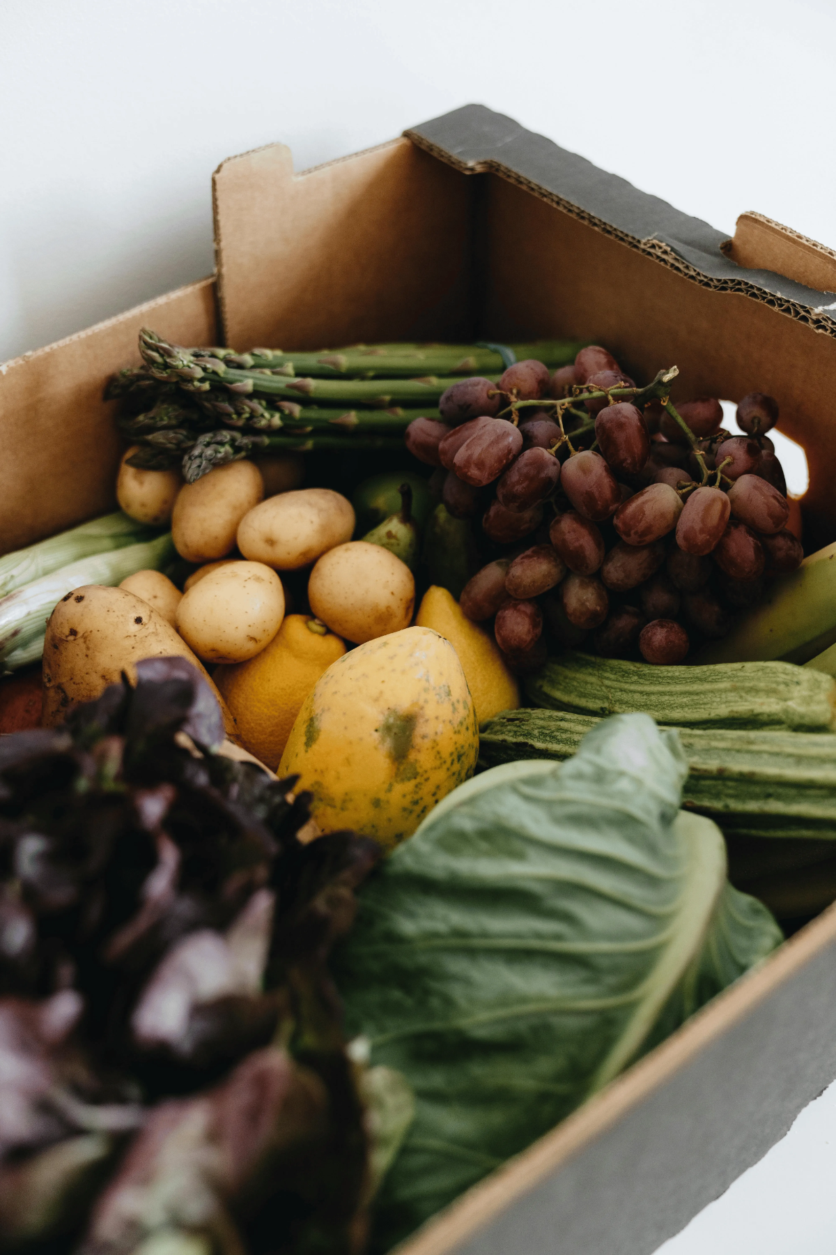 Cardboard delivery box filled with seasonal vegetables and fruit, grapes, asparagus, potatoes, cabbage, squash and more.