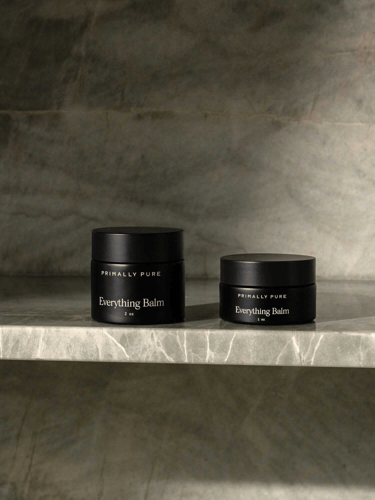 Two black glass jars of Primally Pure “Everything Balm” tallow moisturizer on a marble shelf, 2-oz and 1-oz sizes.