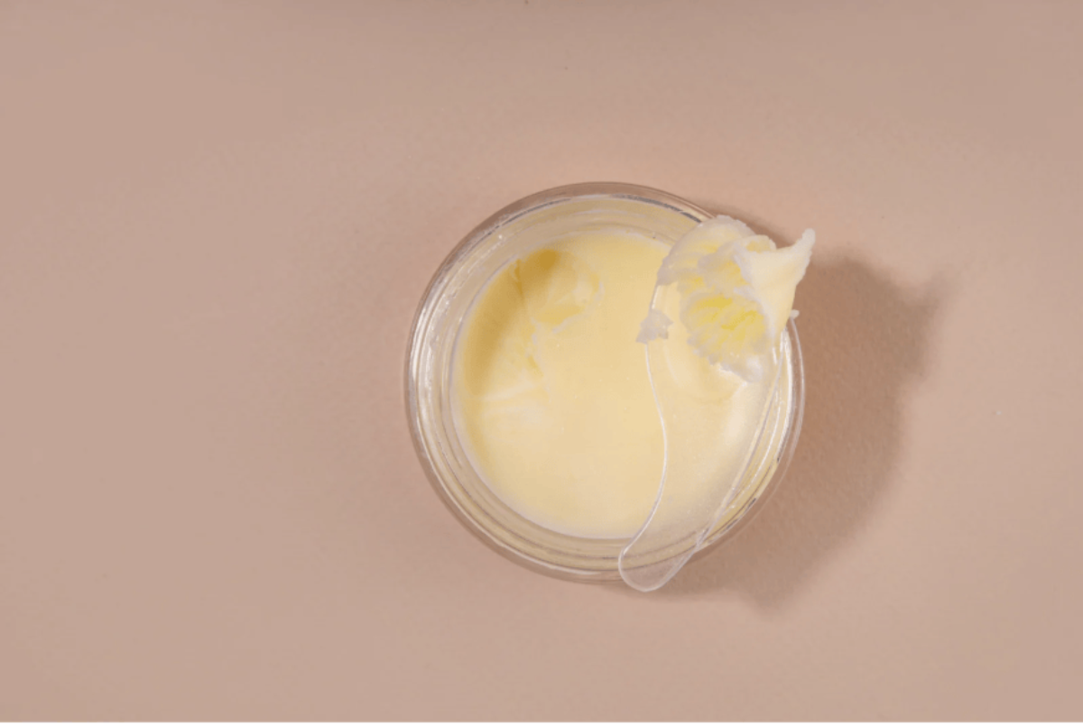Top-view of a glass jar filled with whipped tallow balm, a small scoop resting on the surface, set against a soft beige backdrop.