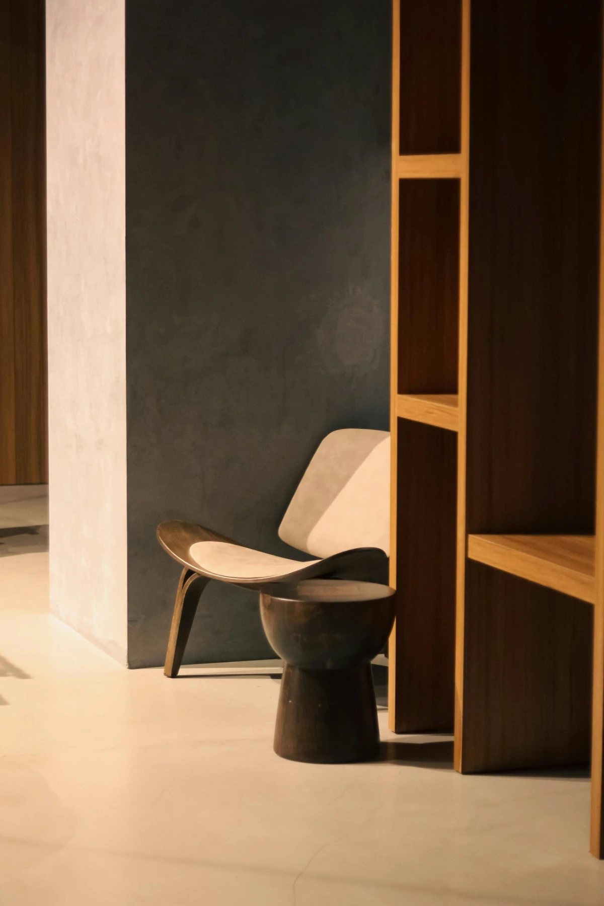 Cozy alcove featuring a low sculptural lounge chair with cream cushion, dark wood stool and built-in shelving.