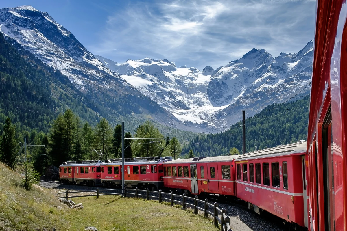 Red alpine train crossing stone bridge through forest with towering snow-covered mountains in the background.