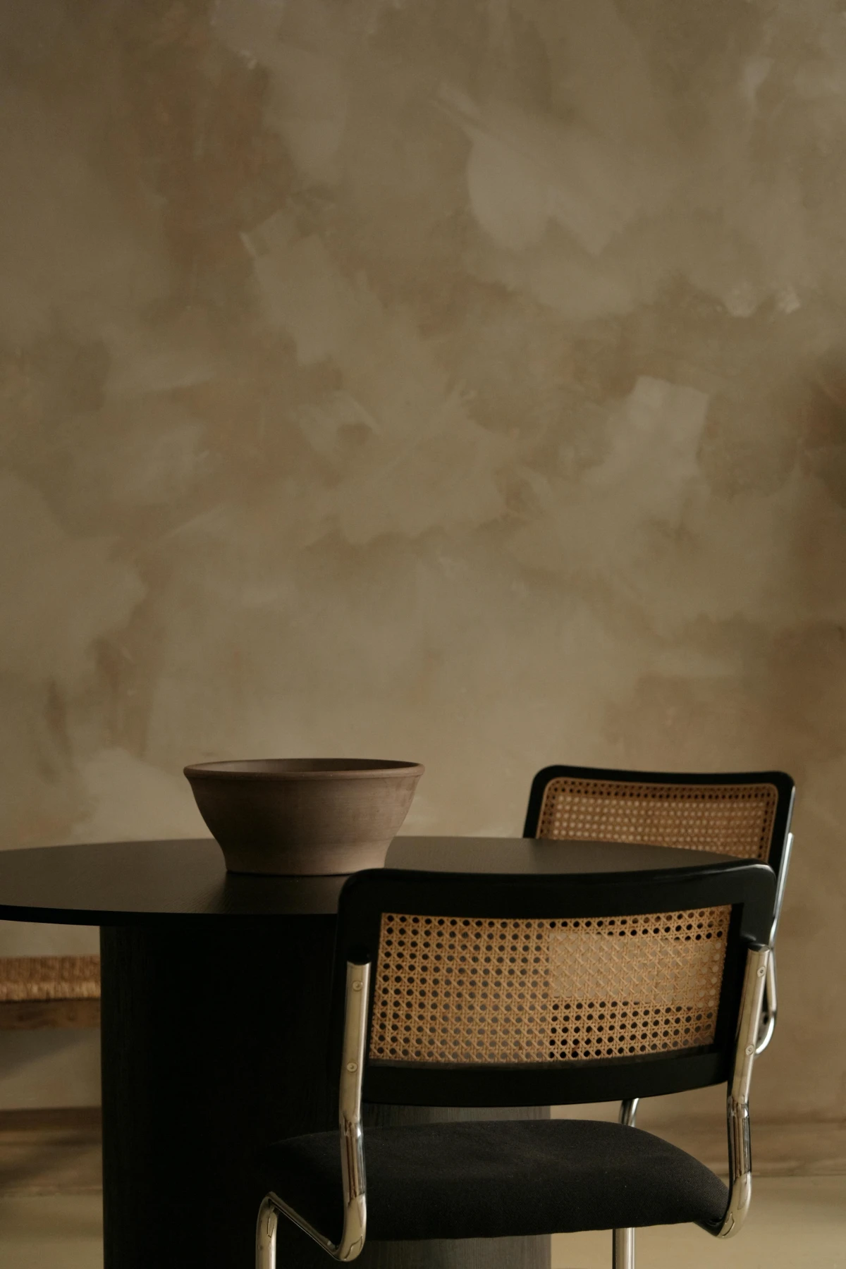 Round black dining table with clay bowl and two black Cesca chairs featuring cane backs, set before a warm plaster wall.