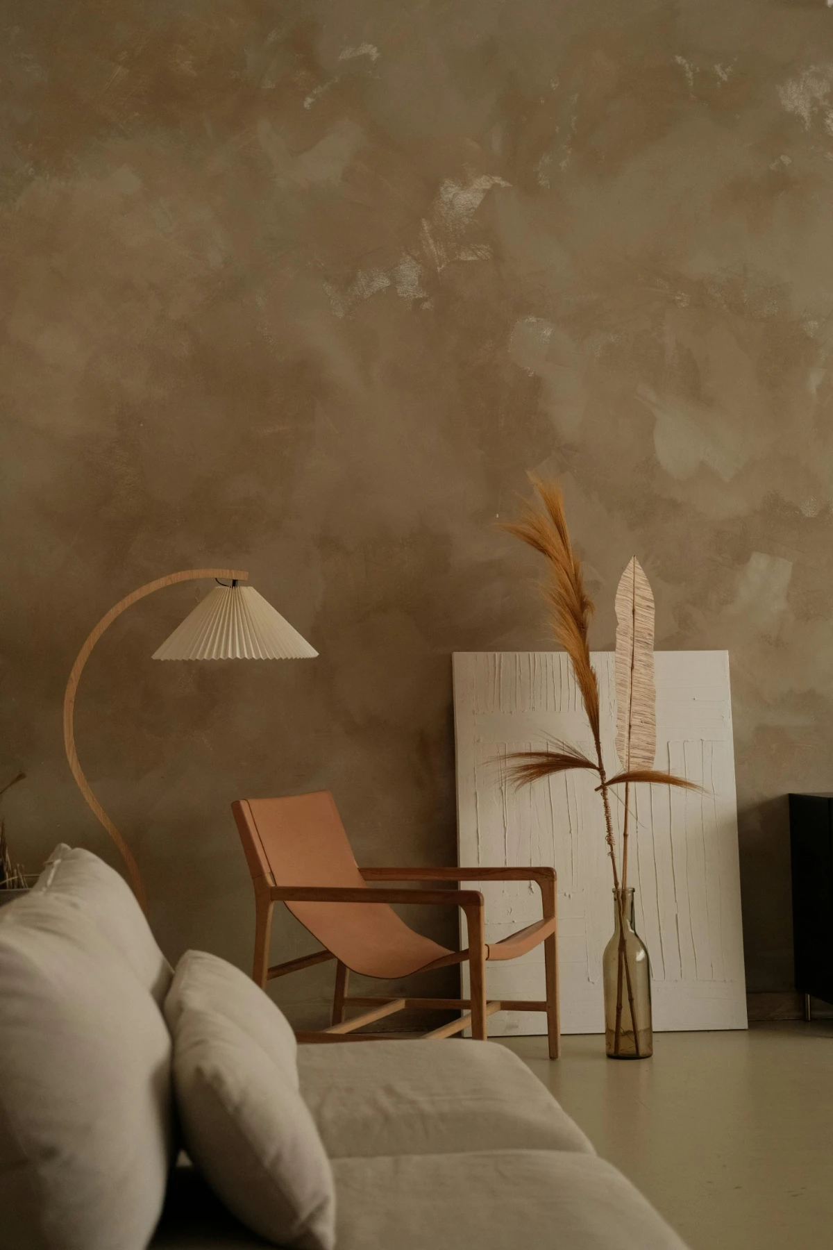 Earth-toned living space with tan leather sling chair, pleated arc floor lamp and dried grasses against a textured khaki wall.