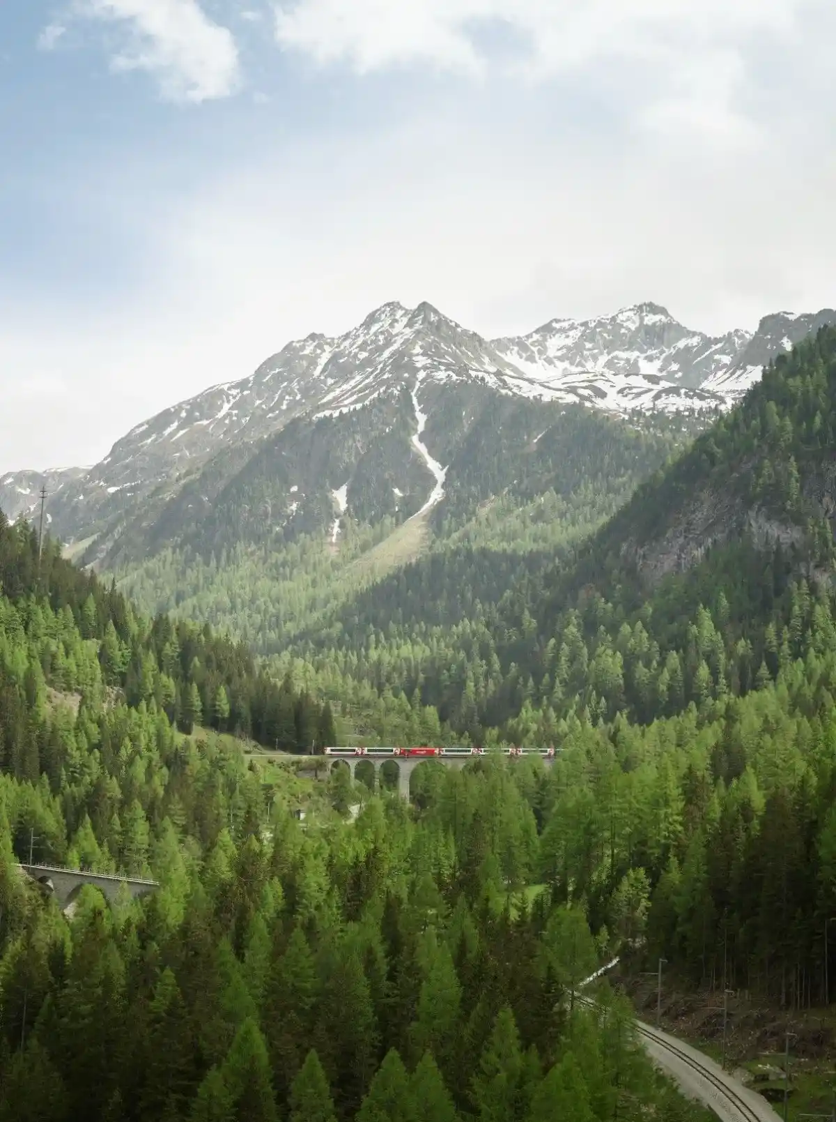 Distant Glacier Express crossing a stone viaduct amid dense evergreen forest and snow-dusted peaks in Graubünden.