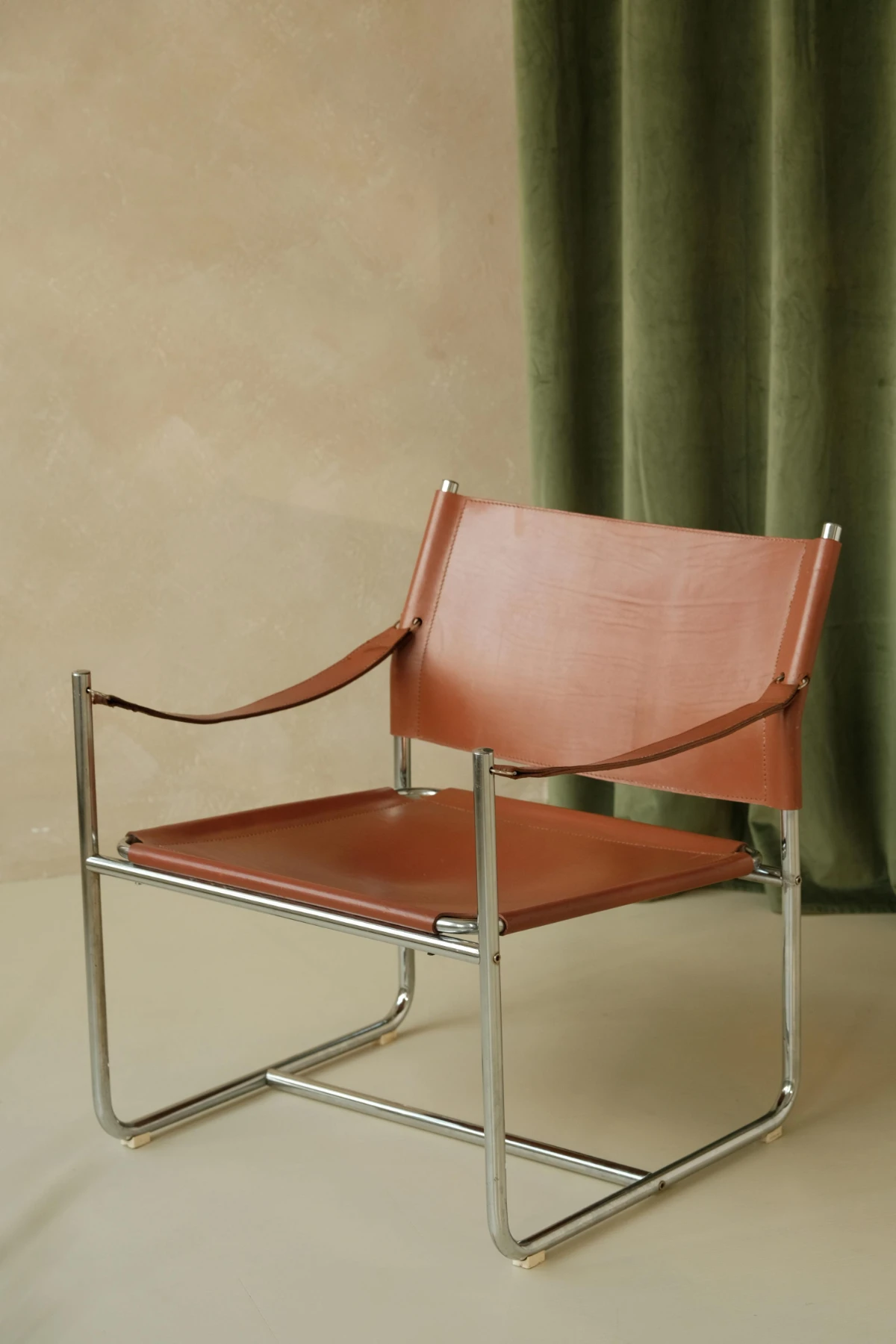 Vintage brown leather sling chair on chrome frame set against a textured wall and green velvet curtain backdrop.
