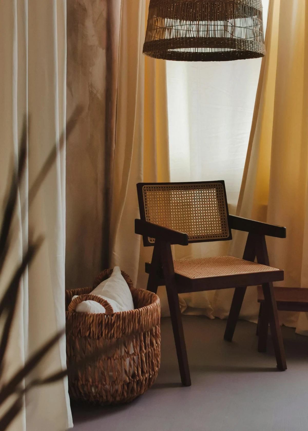 Warmly lit corner with dark wood chair featuring woven rattan seat/back, wicker pendant lamp and basket with cushion.