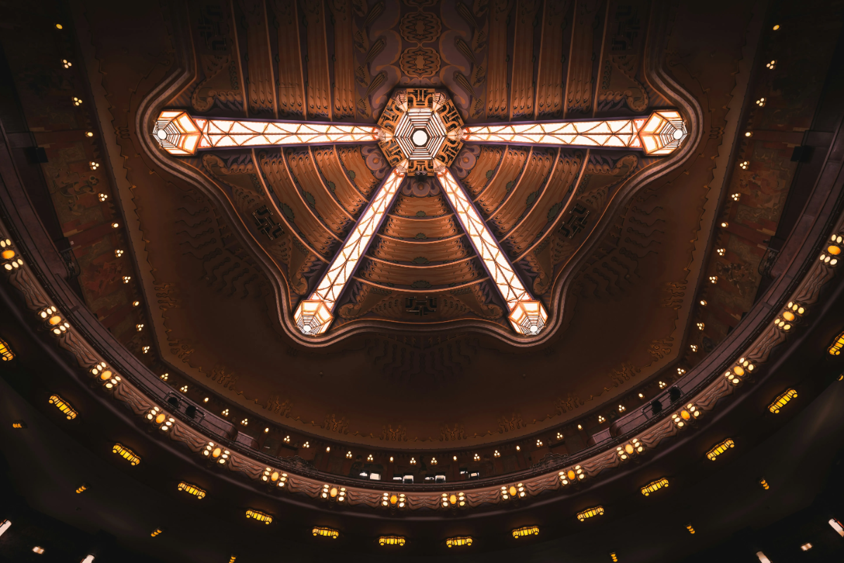 Geometric art-deco chandelier of three illuminated beams radiates from ornate theater ceiling, surrounded by balcony lights.