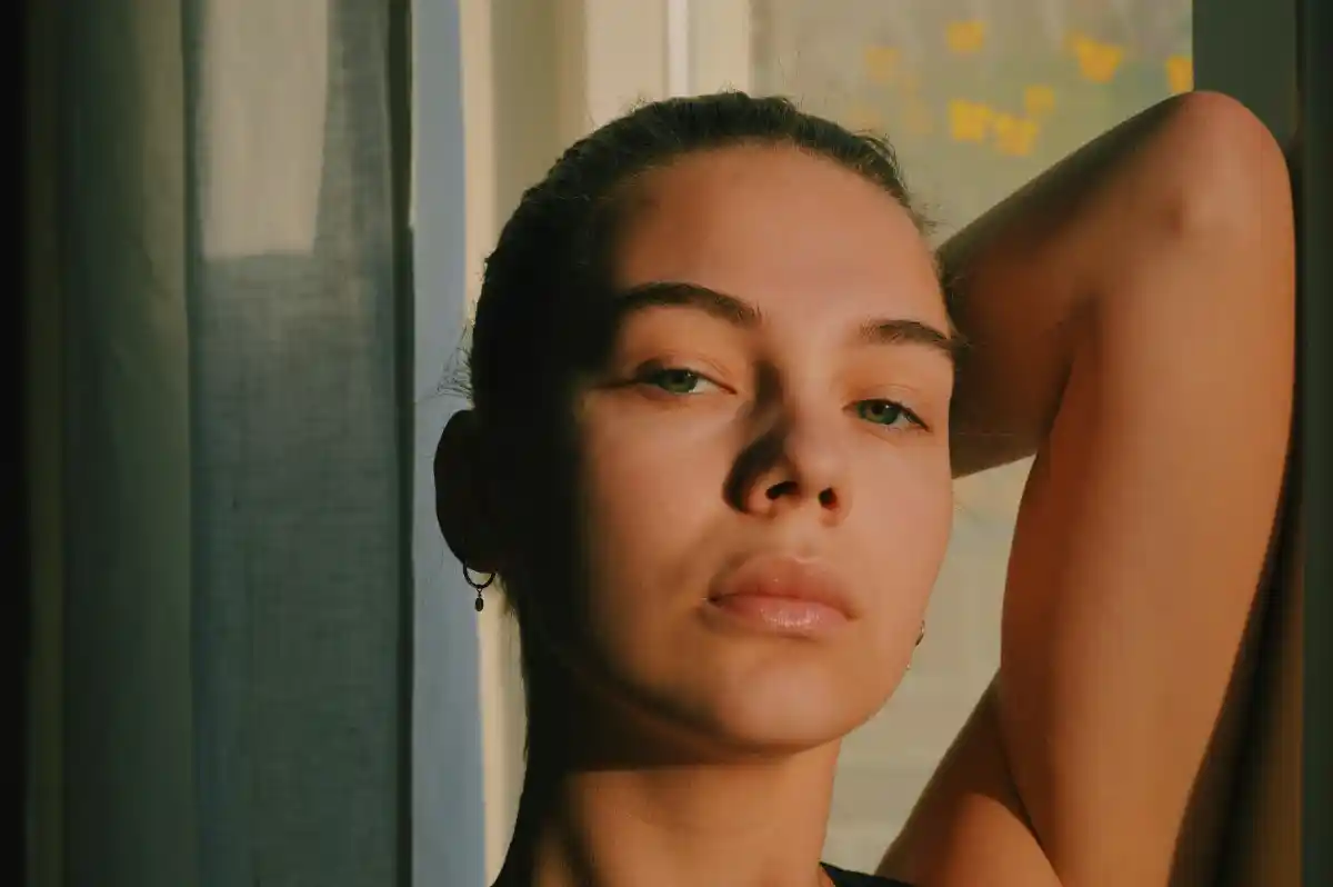 Young woman with bare glowing skin posing in soft window light, arm raised behind head.